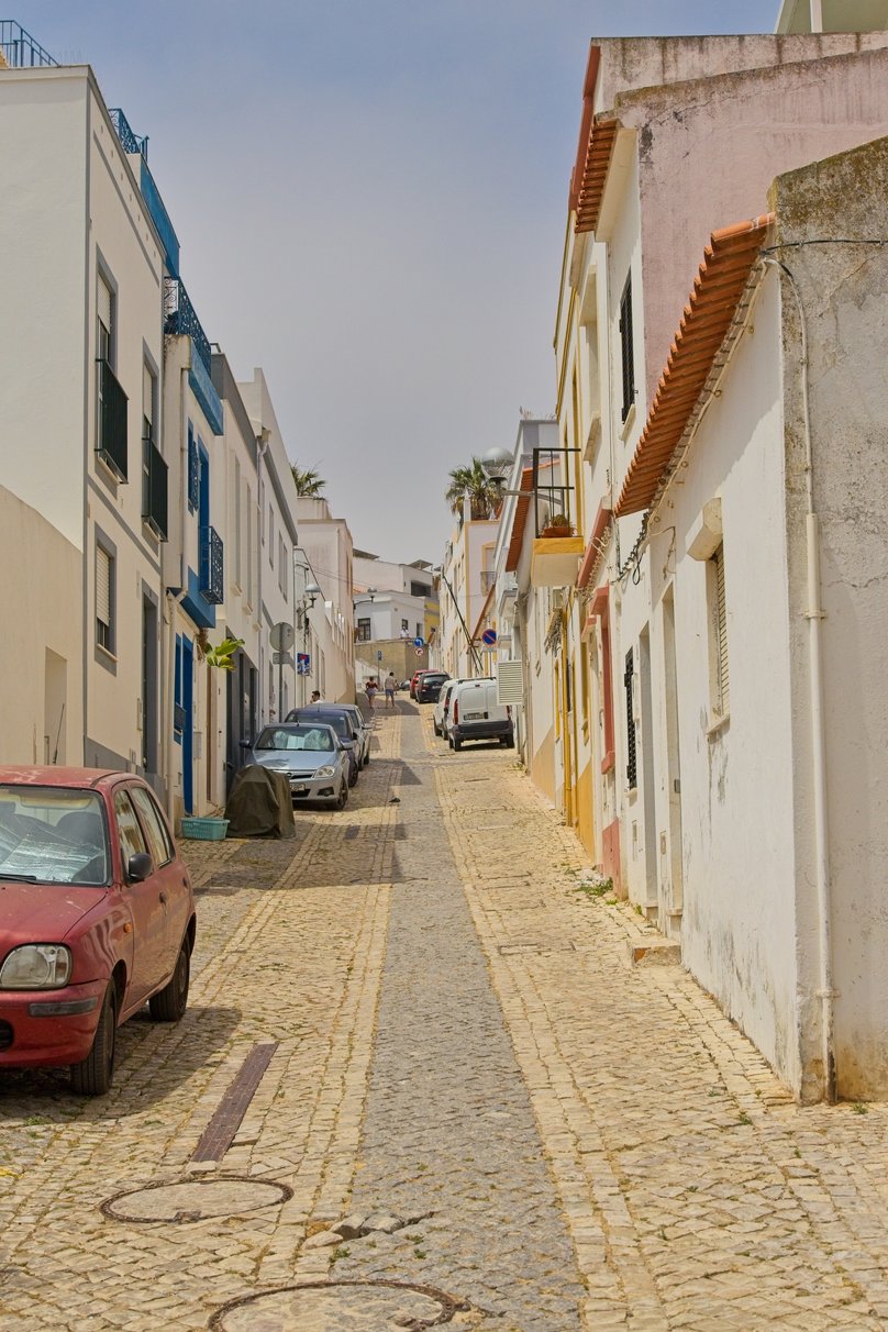 A narrow, gently sloping cobblestone street in a traditional European town, likely Portuguese, lined with closely packed white and pastel-colored buildings. The architecture features flat facades with terracotta roof tiles, blue window frames and balconies, and small planters. Several cars are parked along both sides of the street, including a red car in the foreground and a silver car, with a white van further up. Manhole covers are visible in the stone-paved road. A few pedestrians can be seen walking in the distance under a clear, hazy sky.